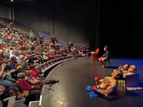 Dans une salle de spectacle, des marionettes sont posées au plateau, une commédienne metteuse en scène répond aux questions du public.