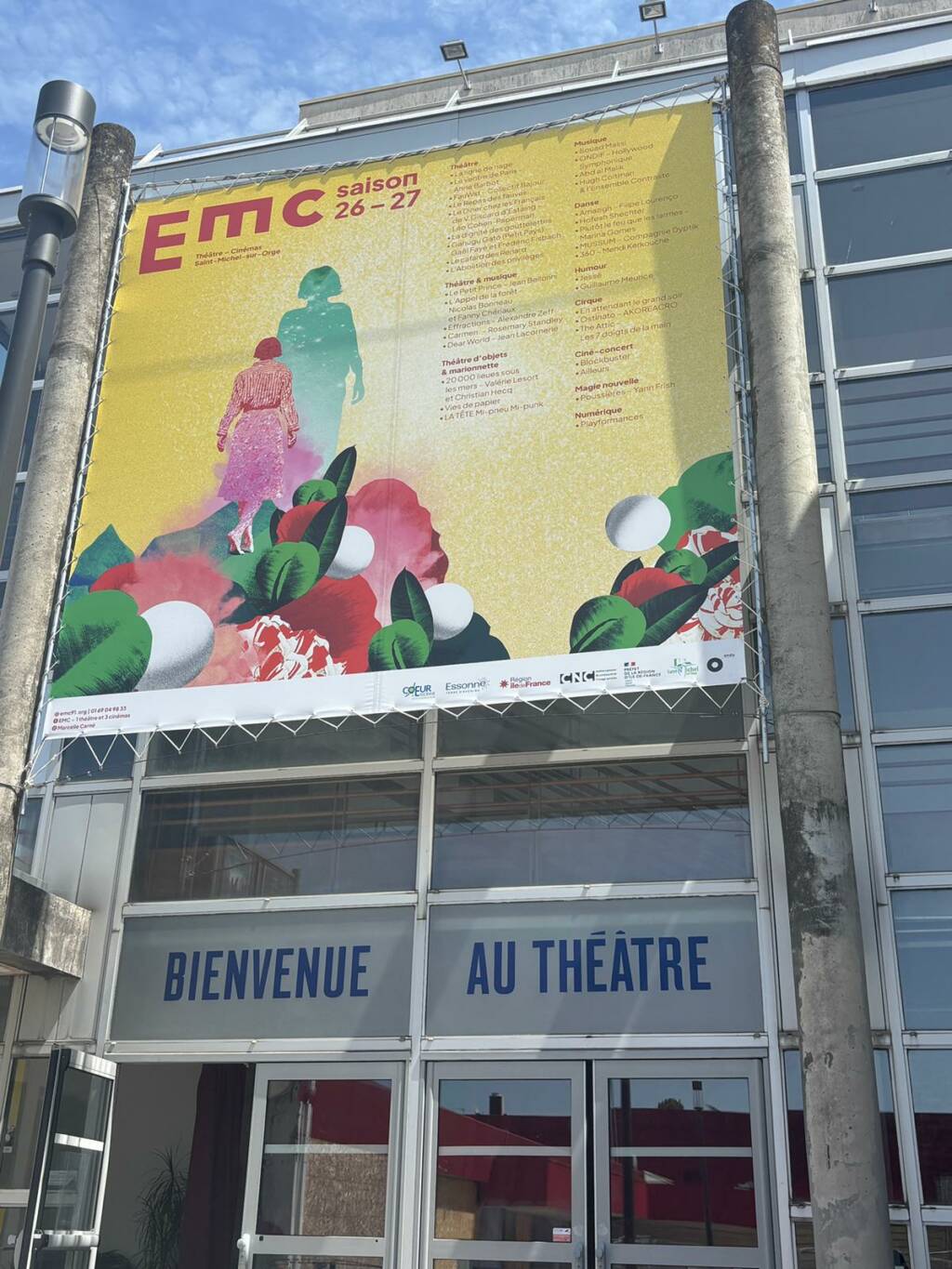 Une bâche sur la façade du bâtiment de l'EMC représente une femme qui marche dans un amas de verdure sur un fond jaune.